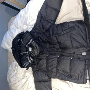 Svart pufferjacka/väst från Burberry storlek M - Svart pufferjacka från Burberry går att göra till en väst också, med stor huva och klassiskt rutigt foder. Jackan har dragkedjor på fickorna, tydlig Burberry-patch på ärmen och quiltad design. Perfekt för kalla dagar och riktigt snygg streetstil.