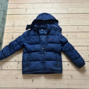 Mörkblå dunjacka från Polo Ralph Lauren - Säljer en mörkblå dunjacka från Polo Ralph Lauren med huva och dragkedja framtill. Jackan har två fickor med dragkedja och klassisk Polo-logga broderad på bröstet. Perfekt för kalla dagar och riktigt snygg passform.