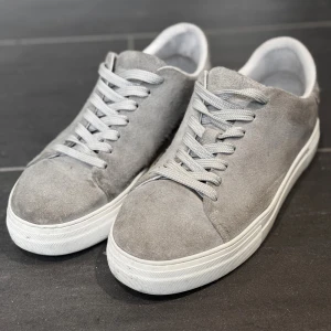 Grå mockasneakers från Selected Homme - Snygga grå sneakers från Selected Homme i mocka med vita sulor och matchande grå snörning. Klassisk låg modell som passar perfekt till jeans eller chinos. Tillverkade i Portugal och har en stilren, minimalistisk look.