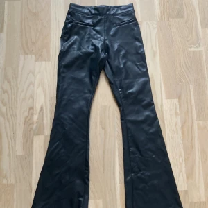 Svarta bootcut byxor i skinnimitation Zara - Snygga svarta bootcut byxor från Zara i skinnimitation. Byxorna har hög midja och en glansig finish som ger en cool look. Perfekta för dig som vill sticka ut med en trendig och edgy stil.
