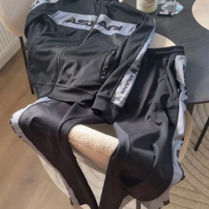 Svarta mjukisbyxor från Askari - Svarta mjukisbyxor från Askarpi med gråa sidopaneler och logga längs benen. Byxorna har resår i midjan och är tillverkade i ett mjukt och stretchigt syntetmaterial. Perfekta för en avslappnad och sportig stil.