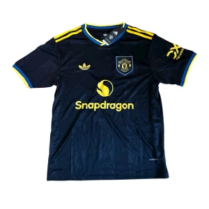 Manchester United x Adidas fotbollströja - Snygg Manchester United fotbollströja i mörkblått med gula detaljer och klubbmärke på bröstet. Adidas-logga och Snapdragon-tryck framtill. Tillverkad i lätt och ventilerande polyester, perfekt för match eller träning.