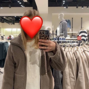 Beige teddyjacka med dragkedja - Mysig beige teddyjacka med dragkedja framtill och hög krage. Jackan har en loose passform och är perfekt för kyliga dagar. Fickor på sidorna och mjukt material som ger en chill vibe.