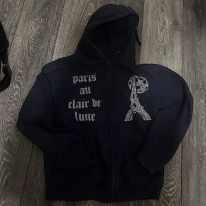 Mörkblå rhinestone blinglucid hoodie  - Mörkblå hoodie med dragkedja och huva. Framsidan har texten 'paris au clair de lune' i strass och en glittrig Eiffeltorn-design med måne. Perfekt statement-plagg för dig som gillar Paris-vibes och snygga detaljer.