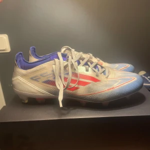 Adidas fotbollsskor blå/röd/vit - Säljer ett par Adidas fotbollsskor med snygg design i vitt, blått och rött. Skorna har snörning, låg profil och mönstrad ovandel i syntetmaterial. Adidas-logga på hälen och klassiska ränder på sidan. Perfekt för dig som vill sticka ut på planen.