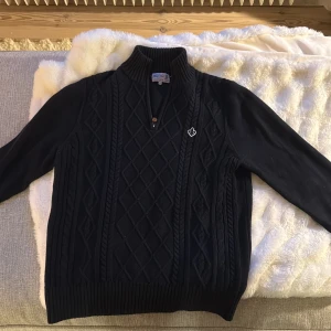 Svart stickad tröja från Morris - Mörkblå stickad tröja från Morris med hög krage och half zip-dragkedja. Snyggt kabelstickat mönster framtill och ribbade muddar. Liten broderad logga på bröstet. Perfekt för kyliga dagar och stilren look.