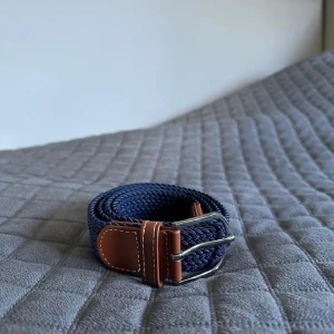 Mörkblå flätat bälte med skinn-detaljer - Snyggt mörkblått flätat bälte med bruna skinn-detaljer och silverfärgat spänne. Bältet har en stilren design med tydliga sömmar på skinnet och passar perfekt till jeans eller chinos. Den flätade strukturen ger en modern och avslappnad känsla.