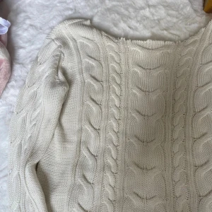 Vit kabelstickad tröja, off shoulder  - Säljer en vit kabelstickad tröja med klassiskt flätmönster. Tröjan har lång ärm, ribbade muddar och är gjord i ett mjukt, stickat material. Perfekt för lager-på-lager och chill höst- eller vinterstil.