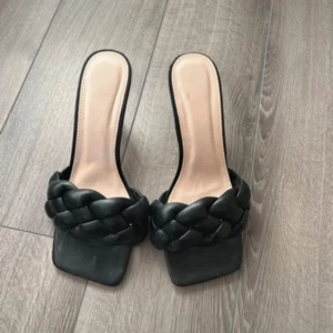 Svarta flätade sandaler med klack - Stilrena svarta sandaler med fyrkantig tå och låg klack. Skorna har en bred, flätad rem i skinnimitation över foten och öppen häl. Perfekta för dig som gillar trendiga och minimalistiska detaljer.