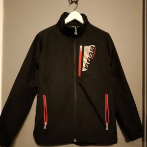 Svart vindjacka från 8848 Altitude - Svart vindjacka från 8848 Altitude med röda dragkedjor och vit logga på bröstet. Jackan har hög krage, meshfoder på insidan och tre fickor med dragkedja. Perfekt för dig som gillar sportig och clean stil.
