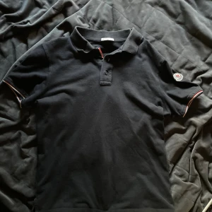 Svart pikétröja från Moncler - Svart pikétröja från Moncler med klassisk krage och två knappar framtill. Ärmarna har snygga rödvita detaljer och Moncler-logga på vänster ärm.                                                                              Pris kan såklart diskuteras✅