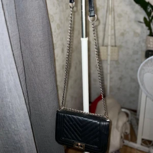 Svart quiltad axelväska med kedja - Snygg svart axelväska i quiltat skinnimitation med silverfärgad kedjerem och vridlås i metall. Väskan har ett klassiskt rutmönster i V-form och ett innerfack med dragkedja. Perfekt storlek för det viktigaste och ger en trendig vibe till din outfit.