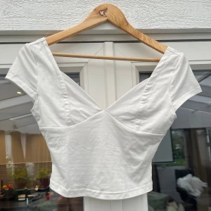 Vit croppad topp med v-ringning - Snygg vit croppad topp med djup v-ringning och korta ärmar. Toppen har en figurnära passform och är sydd i mjuk bomull, perfekt för en trendig och stilren look. Enkel att matcha med både jeans och kjol.