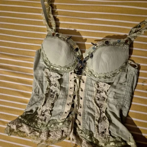 Grön korsett med spets och snörning - Supersöt ljusgrön korsett med spetsdetaljer, volanger och snörning framtill. Dekorerad med små pärlor och kedjor längs bysten. Axelbanden är spetsprydda och hela toppen har romantiska vibbar. Perfekt för dig som gillar unika plagg med mycket detaljer.