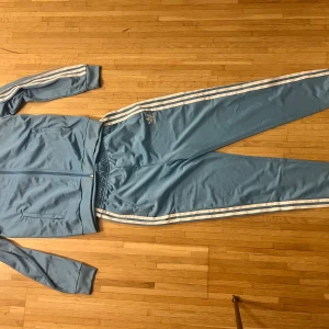 Adidas blå ziptröja med vita ränder retro - Snygg ljusblå ziptröja från Adidas med klassiska vita ränder längs ärmarna och dragkedja framtill. Tröjan har ribbade muddar och två fickor. Perfekt för träning eller chill dagar. Matcha med de tillhörande byxorna för en komplett look.