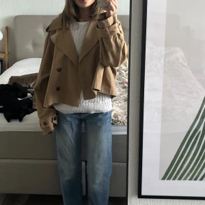 Beige croppad trenchcoat - Säljer en beige croppad trenchcoat med dubbelknäppning, stora slag och breda ärmar med knappar vid ärmslut. Perfekt för dig som gillar en avslappnad men trendig look. Använd som oversized på mig som vanligtvis har stl XS/S i jackor. 