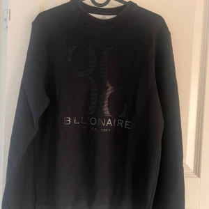 Svart sweatshirt från Billionaire - Svart sweatshirt från Billionaire med tryckt logga och diskret mönster framtill. Tröjan har rund hals, ribbade muddar och är tillverkad i mjuk bomull. Snygg och clean design med exklusiv känsla, perfekt för dig som gillar streetwear med premiumvibe.