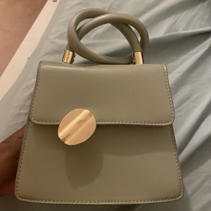 Ljus grön  handväska med gulddetaljer - Stilren beige handväska i syntet med snyggt rundade handtag och guldfärgad rund knäppning framtill. Väskan har en kedja inuti för axelrem och svarta detaljer på insidan. Perfekt för dig som gillar minimalistisk och modern design.