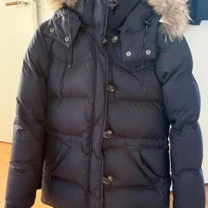 Boomerang Alexandra dunjacka - Mörkblå med avtagbar pälskant på huvan och avtagbar huva. Justerbar midja, vattenavvisande yttertyg. Fylld med 90% RDS certifierat dun och 10% fjäder. Två sidfickor och en innerficka. Passar storlek XXS-S. Nästintill nyskick.