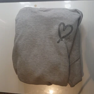 Grå hoodie med hjärta från Unic Studio - Mysig grå hoodie från Unic Studio med stort svart hjärta och texten 'c'est la vie' på bröstet. Tröjan har huva och är tillverkad i mjukt material, perfekt för chill dagar. Enkel och stilren design som passar till det mesta.