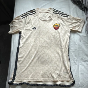 Beige AS Roma fotbollströja Adidas - Säljer en snygg beige AS Roma fotbollströja från Adidas med diskret mönster, klubbmärke på bröstet och klassiska svarta Adidas-ränder på axlarna. Tröjan är kortärmad och tillverkad i lätt och ventilerande material med Aeroready-teknologi.