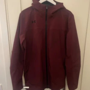 Vinröd vindjacka från Under Armour - Snygg vinröd vindjacka från Under Armour med huva och hel dragkedja framtill. Jackan har en stilren look med svart logga på bröstet och är perfekt för blåsiga dagar. Tillverkad i polyester och har långa ärmar för extra skydd.