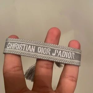 Grått Christian Dior J'Adior armband - Snyggt grått armband från Christian Dior med texten J'Adior broderad i vitt. Armbandet är gjort i vävt tyg och har dekorativa tofsar i ändarna för en trendig look. Perfekt accessoar för att lyfta din outfit.
