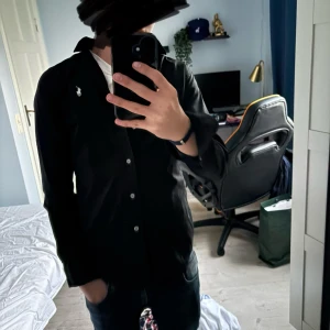 Svart skjorta från Polo Ralph Lauren - Svart skjorta från Polo Ralph Lauren med klassisk krage och vit broderad logga på bröstet. Slim fit-modell med knappar framtill och långa ärmar. Perfekt för dig som gillar stilrena och tidlösa plagg. Skulle uppskatta storleken från 13-14 år