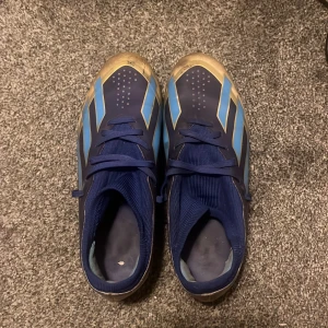 Adidas Messi blå fotbollsskor - Säljer ett par Adidas Messi fotbollsskor i blått med ljusblå och guldiga detaljer. Skorna har snörning och platt sula med dobbar för bra grepp på planen. Snygg design med Messi-tryck på hälen och klassiska Adidas-ränder på sidorna. Perfekt för dig som vill sticka ut på fotbollsplanen.