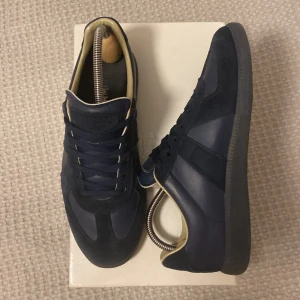 Maison Margiela gats - Säljer ett par stilrena mörkblå sneakers från Maison Margiela med ovandel i både skinn och mocka. Skorna har klassisk snörning, beige innerfoder och en platt sula. Skick 8/10. Storlek 41.