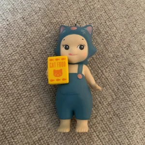 Sonny angel med kattdräkt och kattmat - Söt nyckelring i form av en figur med blå kattdräkt och svans, håller en gul låda med texten 'Cat Food'. Perfekt som dekoration eller present. Figuren är helt ny och inte sliten alls. Pris kan diskuteras!💛