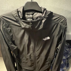 Svart jacka från THE NORTH FACE - Säljer en svart vindjacka från The North Face med luva och dragkedja framtill. Jackan har en clean look med logga på bröstet och ryggen, samt praktiska fickor. Perfekt för blåsiga dagar och enkel att slänga på sig över en hoodie.