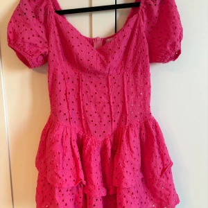 Rosa kortklänning från Lily Rose - Supersöt rosa kortklänning från Lily Rose med broderade hål och volangkjol. Klänningen har puffiga korta ärmar, v-ringning och dragkedja i ryggen. Perfekt för sommarens alla tillfällen när du vill sticka ut lite extra! Har shorts inbyggda. 