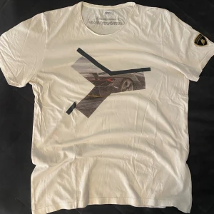 Vit Lamborghini t-shirt med tryck - Vit t-shirt från Lamborghini med coolt grafiskt tryck av en sportbil på bröstet och svart Lamborghini-logga på ryggen. På vänster ärm sitter ett broderat märke i svart och guld. Rund hals och mjukt bomullsmaterial, perfekt för bilentusiasten. Storlek XL men passar L