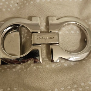 Svart Ferragamo bälte med silverdetalj - Snyggt svart bälte från Ferragamo med ikoniskt, glänsande silverspänne i metall. Bältet är gjort i äkta skinn och har en stilren, klassisk design som lyfter vilken outfit som helst. Perfekt accessoar för dig som gillar exklusiva detaljer och tidlös stil.