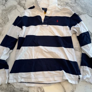 Långärmad piké från Polo Ralph Lauren - Klassisk långärmad pikétröja från Polo Ralph Lauren i bomull. Storlek 14-16 år. Tröjan har breda marinblå och vita ränder, den ikoniska röda loggan broderad på bröstet. Perfekt för en causal och stilren look.