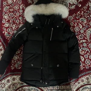 Svart Moose Knuckles dunjacka med päls - Säljer en svart dunjacka från Moose Knuckles med stor vit päls runt huvan. Jackan har flera dragkedjor, både på ärmar och fram, samt snygga detaljer och en quiltad look. Perfekt för kalla dagar och riktigt stilren.
