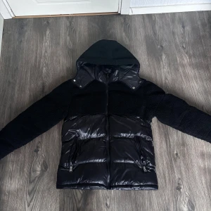 Svart pufferjacka med teddy från boohooMAN - Svart pufferjacka från boohooMAN i storlek S. Jackan har en kombination av glansigt puffer-material och mjuka teddy-detaljer på ärmar, axlar och huva. Utrustad med dragkedja framtill och två fickor med dragkedja. Perfekt för kalla dagar med sin feta look. 