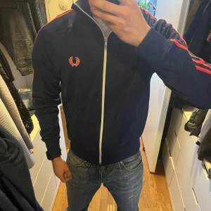 Mörkblå Fred Perry tröja - Säljer en mörkblå track jacket från Fred Perry i storlek S. Jackan har röda ränder längs ärmarna, vit dragkedja och Fred Perry-logga på bröstet. Perfekt för dig som gillar streetwear. Skriv om du har några funderingar!😁