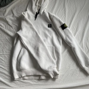 Vit hoodie från Stone Island - Vit hoodie från Stone Island med klassisk patch på ärmen och svart logga på bröstet. Tröjan har huva med snörning, känguruficka och ribbade muddar. Perfekt för dig som gillar stilrena och exklusiva streetwear-plagg.