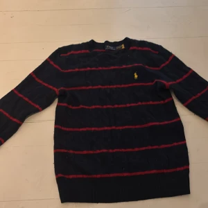 Randig tröja från Polo Ralph Lauren XS - Mörkblå stickad tröja från Polo Ralph Lauren i storlek XS. Tröjan har röda horisontella ränder och den klassiska gula loggan broderad på bröstet. Långärmad och med ribbade muddar för en snygg passform.