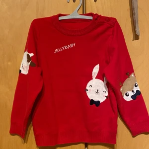 Barntröja, barnkläder, Röd stickad tröja Jellybaby med djurmotiv - Supergullig röd stickad tjock tröja från Jellybaby med broderade djurmotiv på magen och ärmarna. Tröjan har rund halsringning, knappar på axeln och långa ärmar. Perfekt för dig som gillar färgglada och lekfulla plagg!