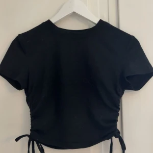 Svart croppad topp med knyt - En svart croppad topp med korta ärmar och snygga knytband i sidorna. Toppen har en enkel och stilren design som passar perfekt till jeans eller kjol. Materialet känns mjukt och bekvämt mot huden.