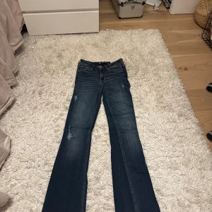 Mörkblå bootcut jeans Hollister W23L - Snygga mörkblå bootcut jeans från Hollister med klassisk femficksdesign och guldfärgad logga på bakfickan. Jeansen har midwaist, lätt slitna detaljer och är tillverkade i mjuk bomullsdenim med stretch. Perfekta för dig som gillar en tidlös och avslappnad look.