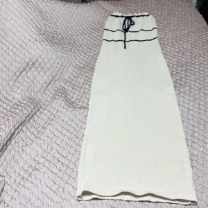 Krämvit klänning  - Krämvit klänning med svart snörning i midjan och två svarta linjer som detalj upptill. Kjolen har rak passform och är enkel men ändå stilren, perfekt för dig som gillar minimalistisk stil.