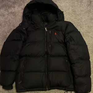 Svart dunjacka från Polo Ralph Lauren - Säljer en svart dunjacka från Polo Ralph Lauren med huva och dragkedja. Jackan har klassisk pufferstil, flera fickor med dragkedja och den ikoniska röda Polo-loggan broderad på bröstet. Perfekt för kalla dagar och riktigt snygg streetstil. En perfekt jacka inför hösten, Kvitto finns✅priset är ej hugget i sten!