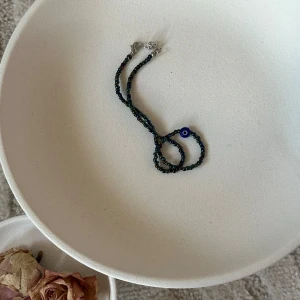 Halsband med blått öga och pärlor - Unikt halsband med små glansiga pärlor i mörka nyanser och ett blått evil eye-berlock i mitten. Perfekt accessoar för dig som gillar detaljer och vill sticka ut med en personlig touch.