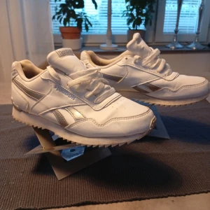 Vita Reebok sneakers med silverdetaljer - Snygga vita Reebok sneakers med klassisk siluett och silverfärgade detaljer på sidorna. Skorna har snörning, rund tå och platt sula med vågigt mönster undertill. Ovandelen är i skinn och de har en stilren look som passar till det mesta.