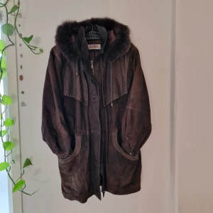 Läderjacka med päls - Fantastisk kappa I mjukt läder(mocka?!)  med päls i kragen. 2 fickor fram, 1 innerficka, dragsko i midjan och huva. Fantastisk under hösten och vintern med några lager under. Syns att den är använd bak vid rumpan, men inget som syns i vardagen tycker jag (se bild). Pga flyttstäd behöver denna hitta ett nytt hem. Mått: Ärm - 60 cm, Axelbredd - 48 cm, Längd - 85 cm. 
