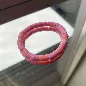 Rosa armband med platta pärlor - Fint armband med platta pärlor i olika nyanser av rosa och lite orange. Armbandet har en rund form och är elastiskt, vilket gör det enkelt att ta på och av. Perfekt accessoar för att addera färg till din outfit.